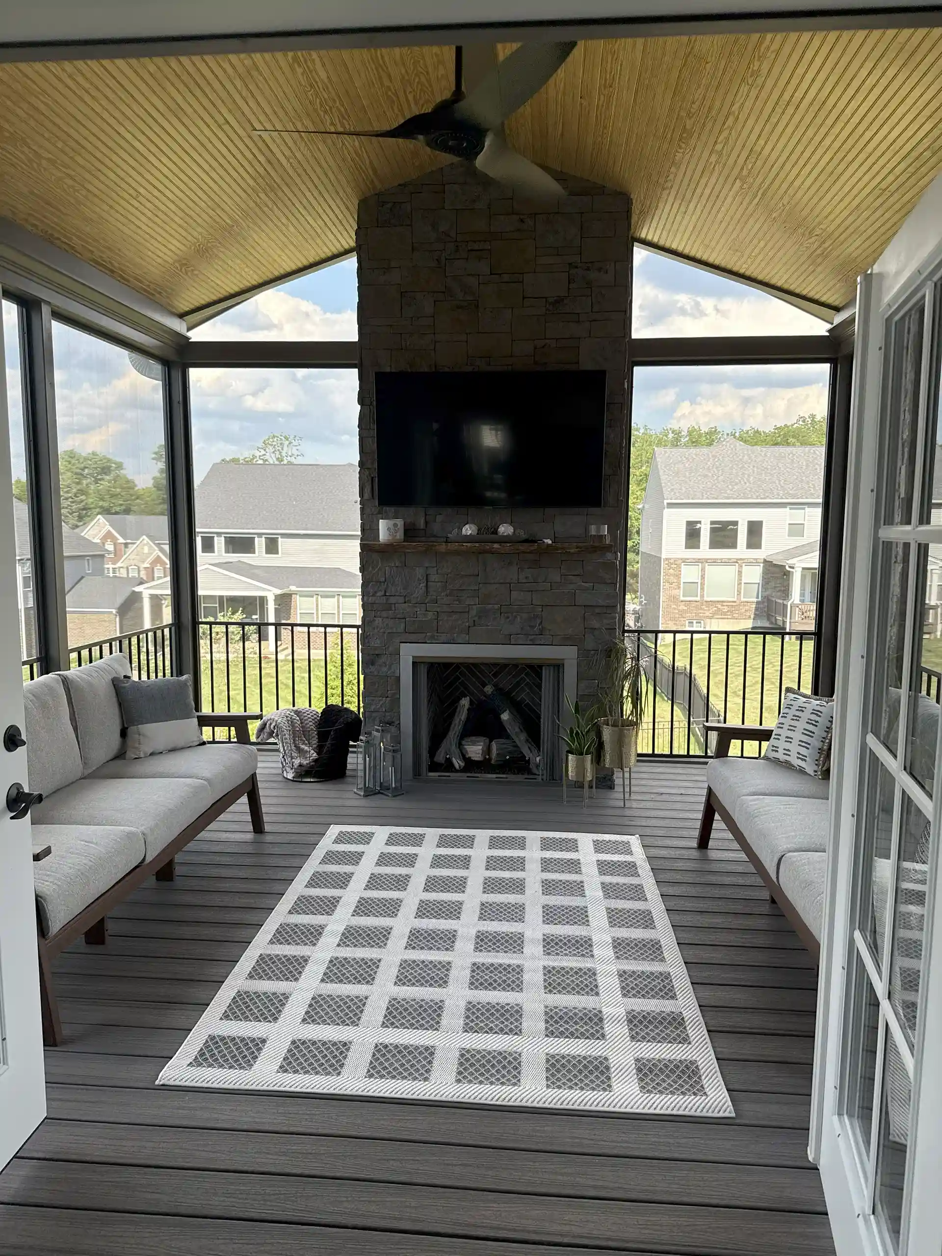 Screened In Porch Cost Guide 2025 (Full Price Breakdown) 1 A screened porch interior with a stone fireplace, seating set, decorative rug, and panoramic windows overlooking a residential neighborhood.