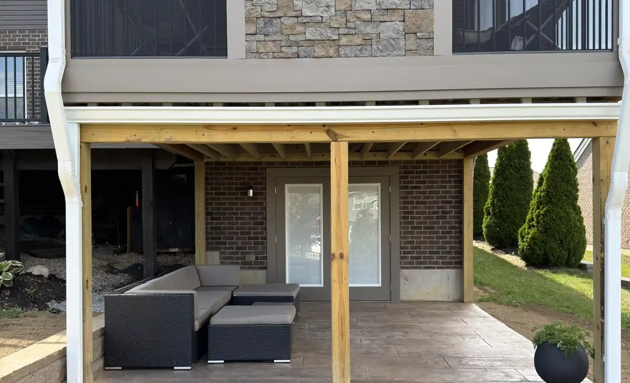 A two-story outdoor living area featuring a lower covered patio with modern seating and a set of double doors, beneath an upper screened-in porch with a central stone wall and black aluminum railing.