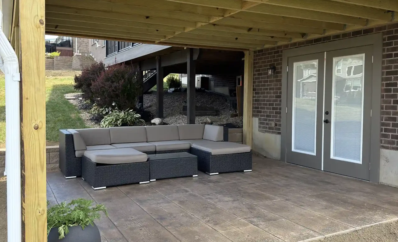 View of a lower covered patio beneath a screened-in porch, featuring modern sectional seating, a potted plant, and double doors leading to the backyard.