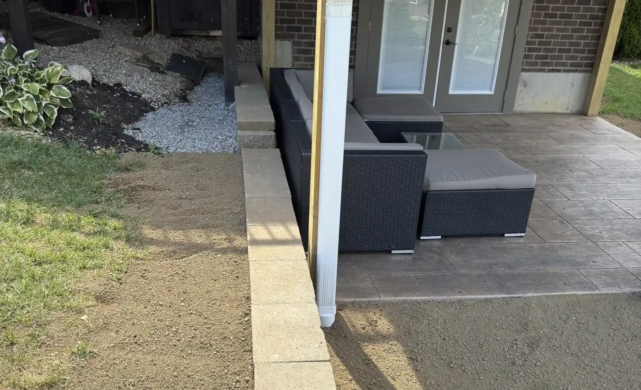 Side view of a covered patio beneath a screened-in porch, showing a stone retaining wall, stamped concrete floor, and double glass doors leading to the backyard.