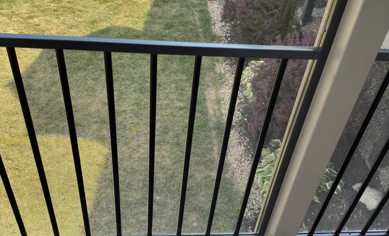 View from inside a screened-in porch, showing a black aluminum railing, TREX composite decking, and a grassy backyard with landscaping.