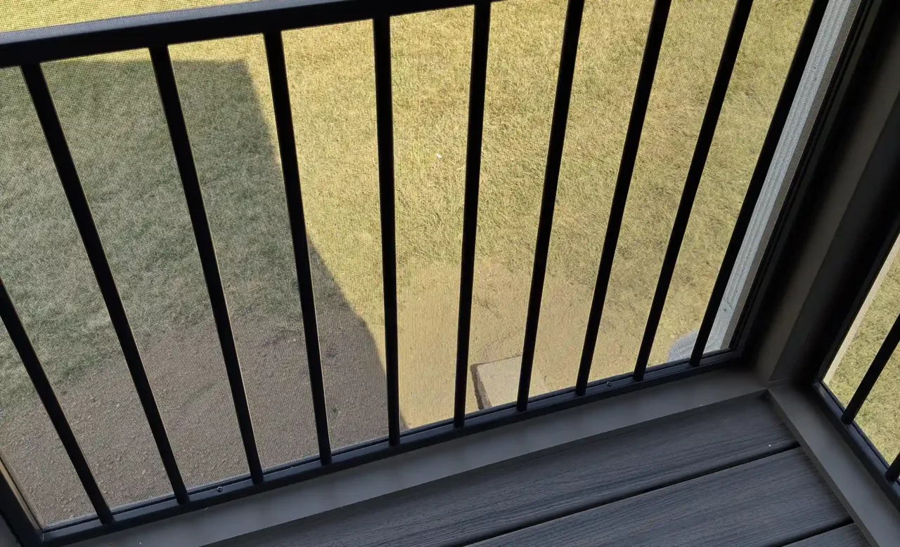 View from inside a screened-in porch, showing a black aluminum railing, TREX composite decking, and a grassy backyard with landscaping.
