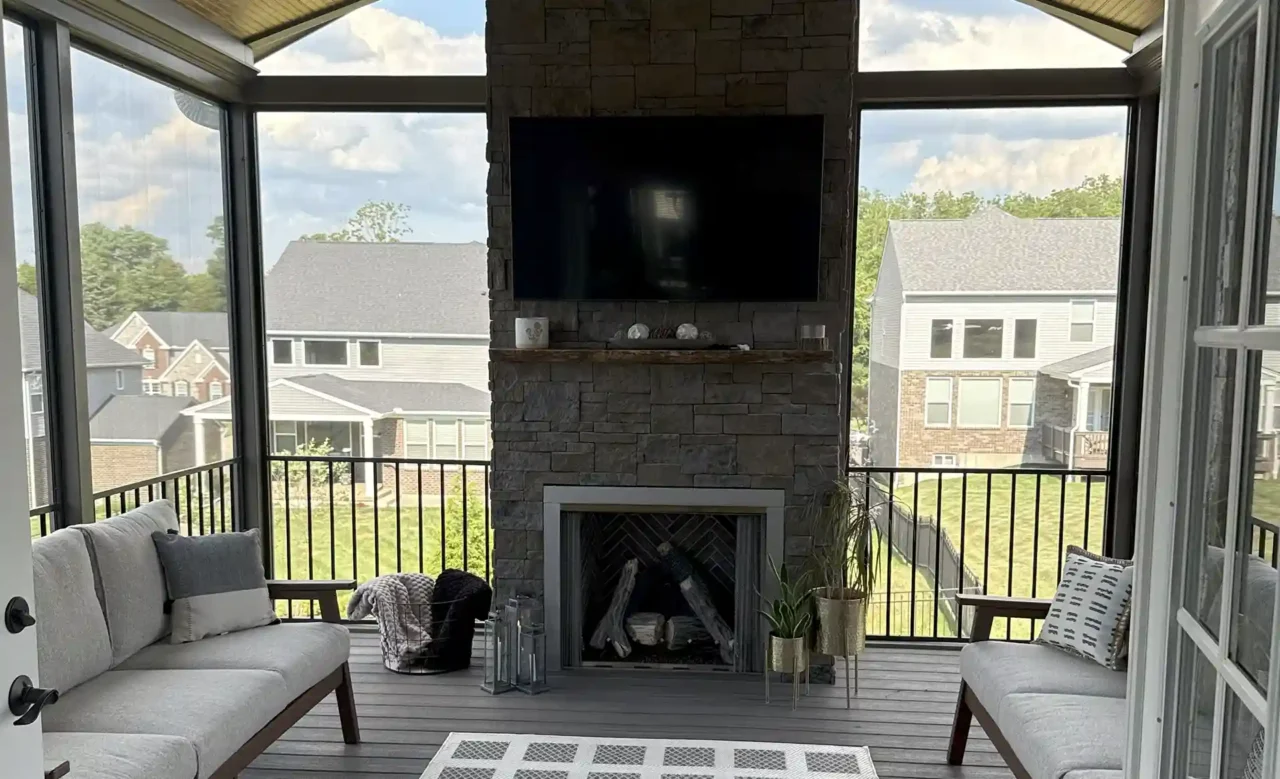 A screened-in porch featuring a stone fireplace, TV mounted above, comfortable seating, and a modern rug, with large windows