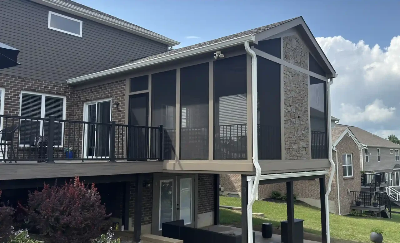 Angled view of a screened-in porch with stone accents and black aluminum railing, featuring large screened panels. Beneath the porch, a dry walk-out patio