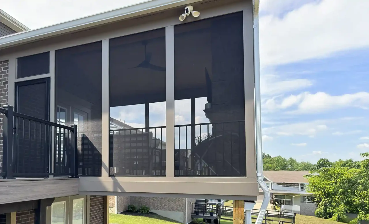 Angled view of a screened-in porch with black aluminum railing and a ceiling fan, featuring large screened panels. Beneath the porch, wooden support beams are visible, along with a dry walkout patio and outdoor seating area.