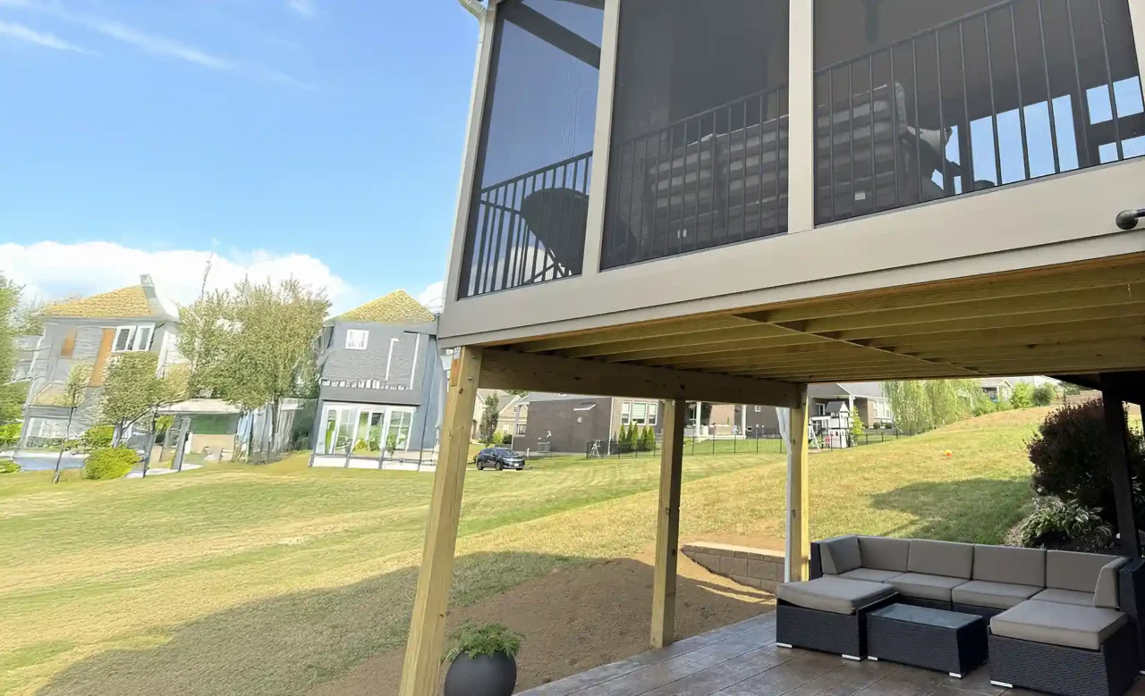 View of a screened-in porch with black aluminum railing and a ceiling fan, featuring large screened panels. Beneath the porch, wooden support beams are visible, with a dry walk-out patio and sectional seating.