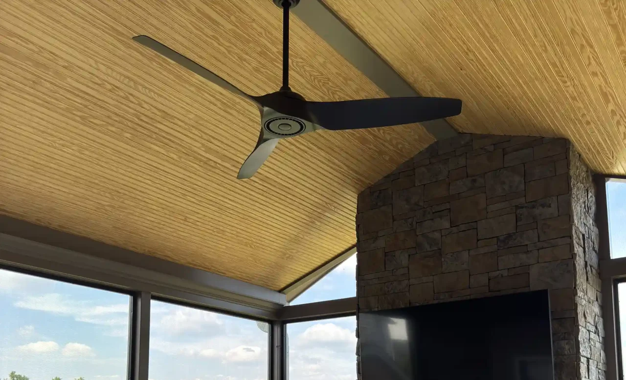 View from the right of a screened-in porch featuring a stone fireplace with a TV mounted above, a modern rug, seating, and a ceiling fan, with large windows