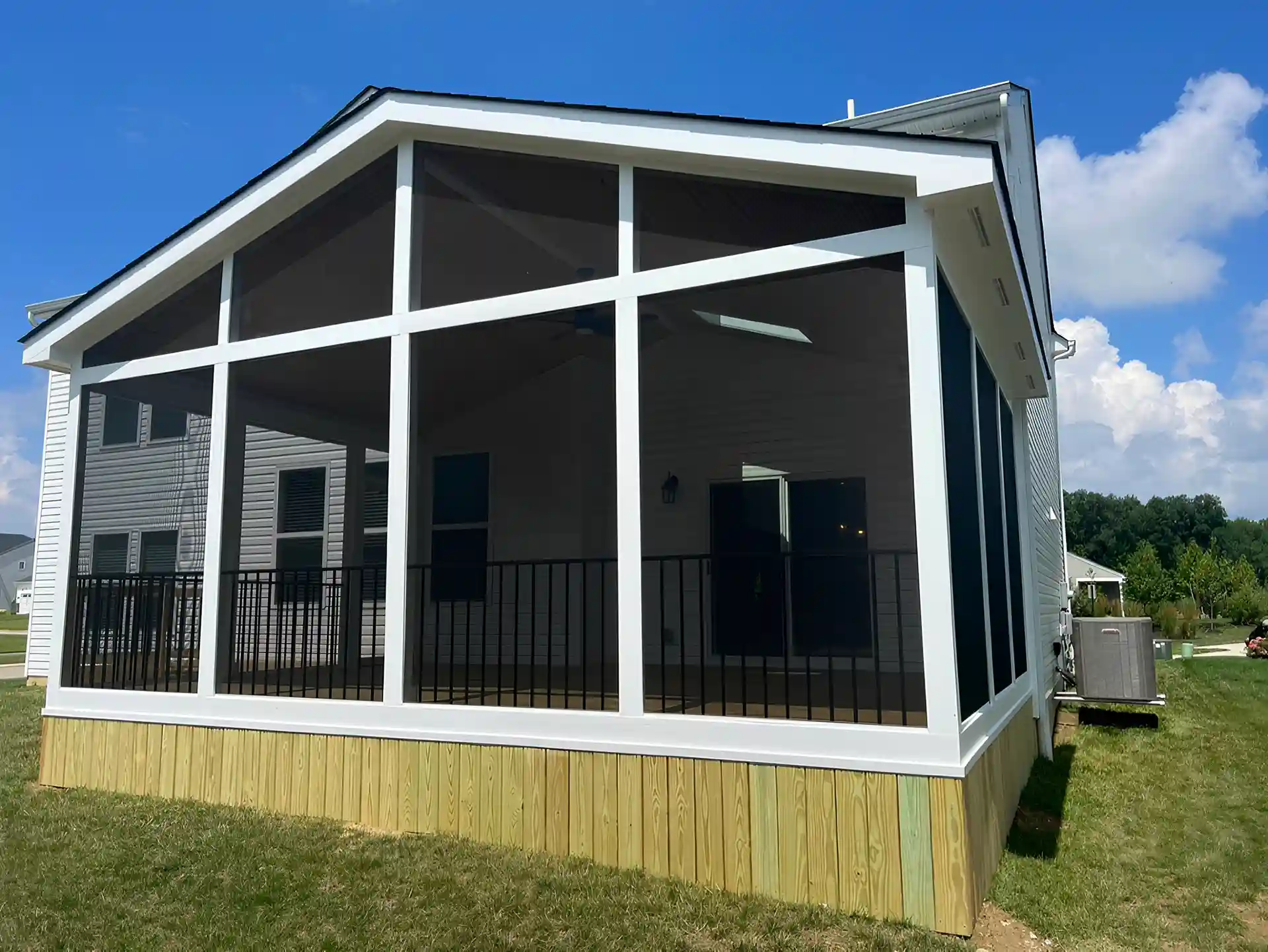 Screened porch with gable roof, black railing, and wood skirting attached to house.