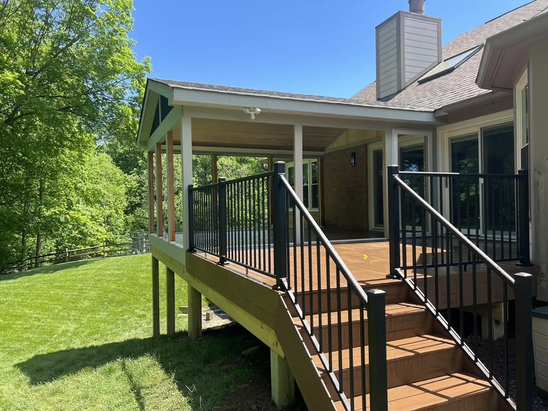 Best Time to Build a Deck A composite deck extends from a two-story house with tan siding and a dark brown roof. The deck features a covered porch area and a set of stairs leading down to a grassy backyard. The deck and stairs have a composite brown finish and are secured by black metal railings.