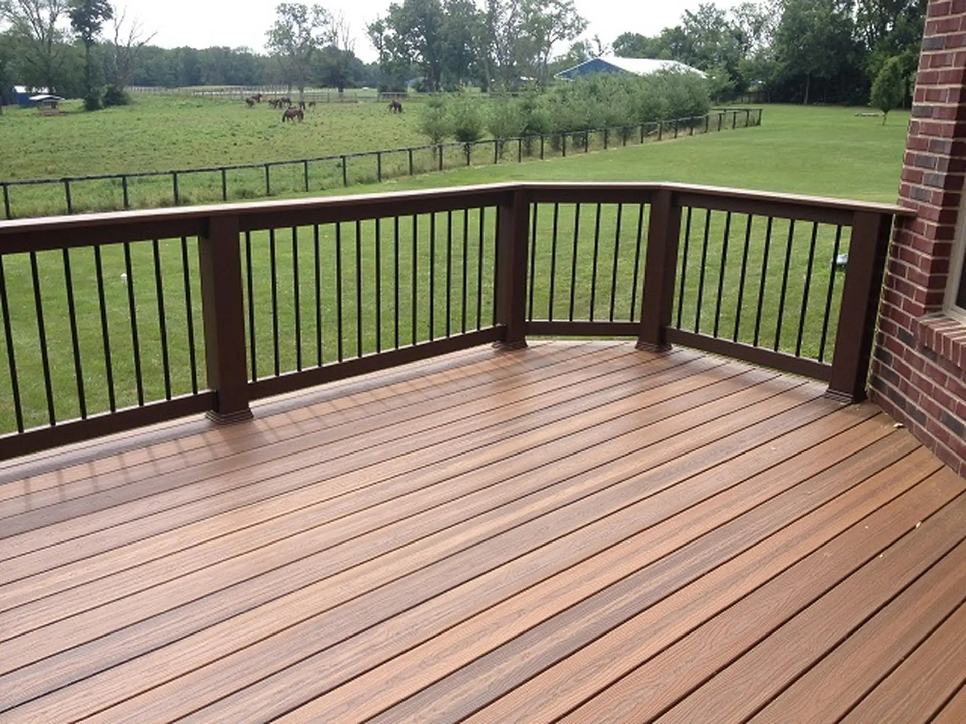 When is the Best Time to Build a Deck A corner section of a wooden deck with a dark brown railing and black metal balusters. The decking is composed of reddish-brown planks. The deck is next to a red brick exterior wall of a building.