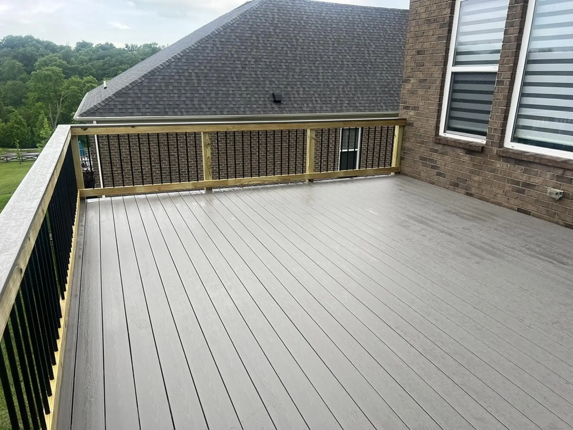 An outdoor composite deck with gray deck boards. The deck has a railing system with black metal balusters and a wood top rail. The deck is attached to a house with a reddish-brown brick exterior and windows with horizontal blinds. In the background, a large house with a dark gray shingled roof is visible.