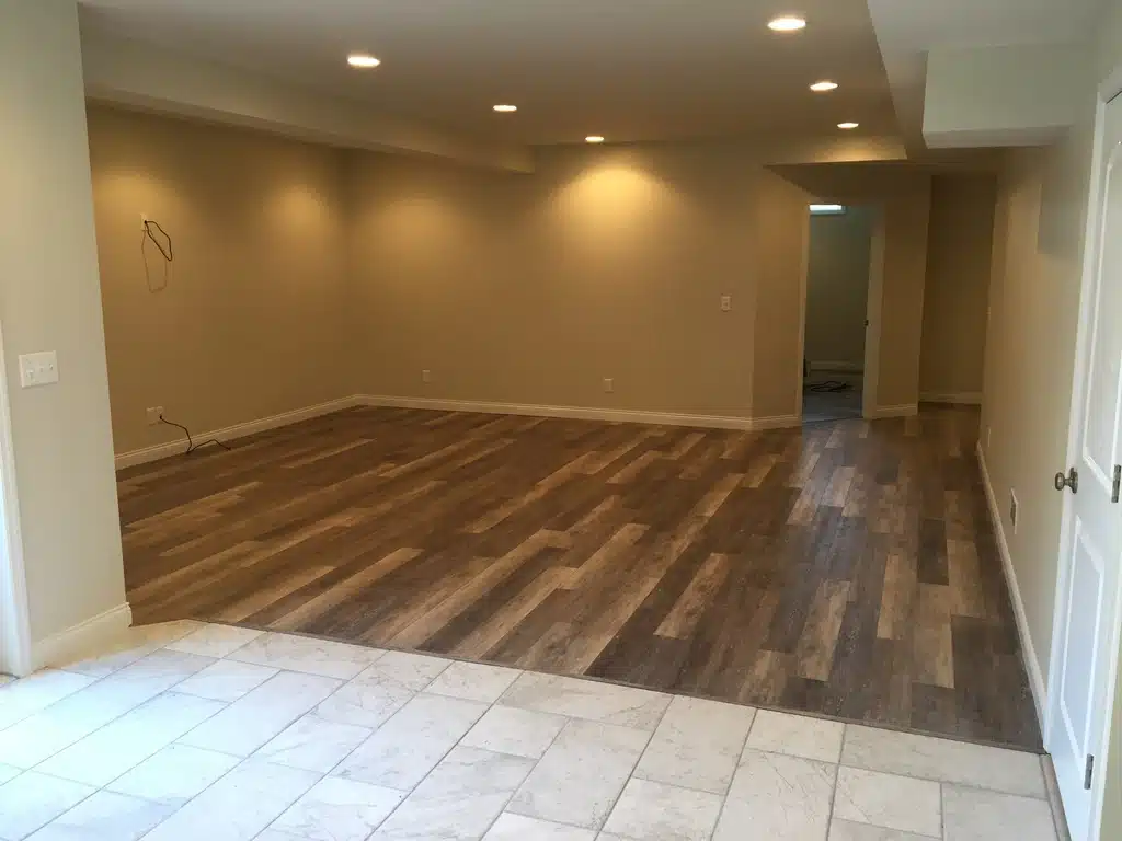 A spacious, empty room featuring a combination of flooring types and warm lighting. The foreground consists of light-colored, square tiles that transition into a larger area of dark wood-look laminate or vinyl plank flooring. The walls are painted in a neutral beige or tan tone, and the ceiling is equipped with several recessed "can" lights that illuminate the space. 