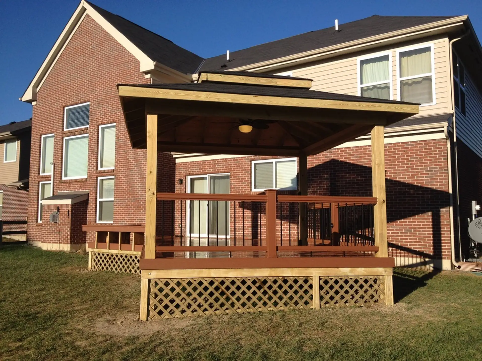 Covered wooden pavilion on elevated deck with lattice skirting, wood posts, shingled roof and ceiling fan next to a two-story brick and siding house