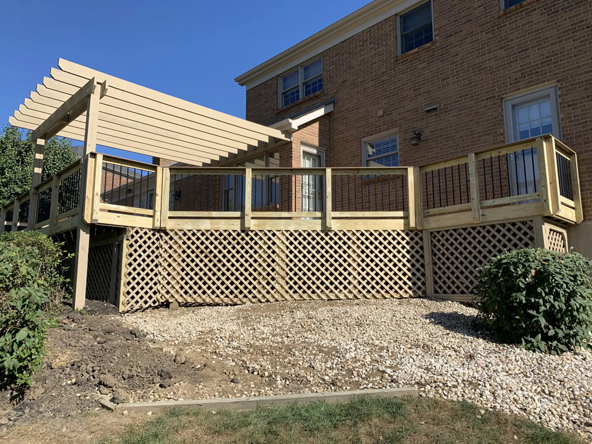 Raised wooden deck attached to a brick house with a pergola, lattice skirting and black metal baluster railing