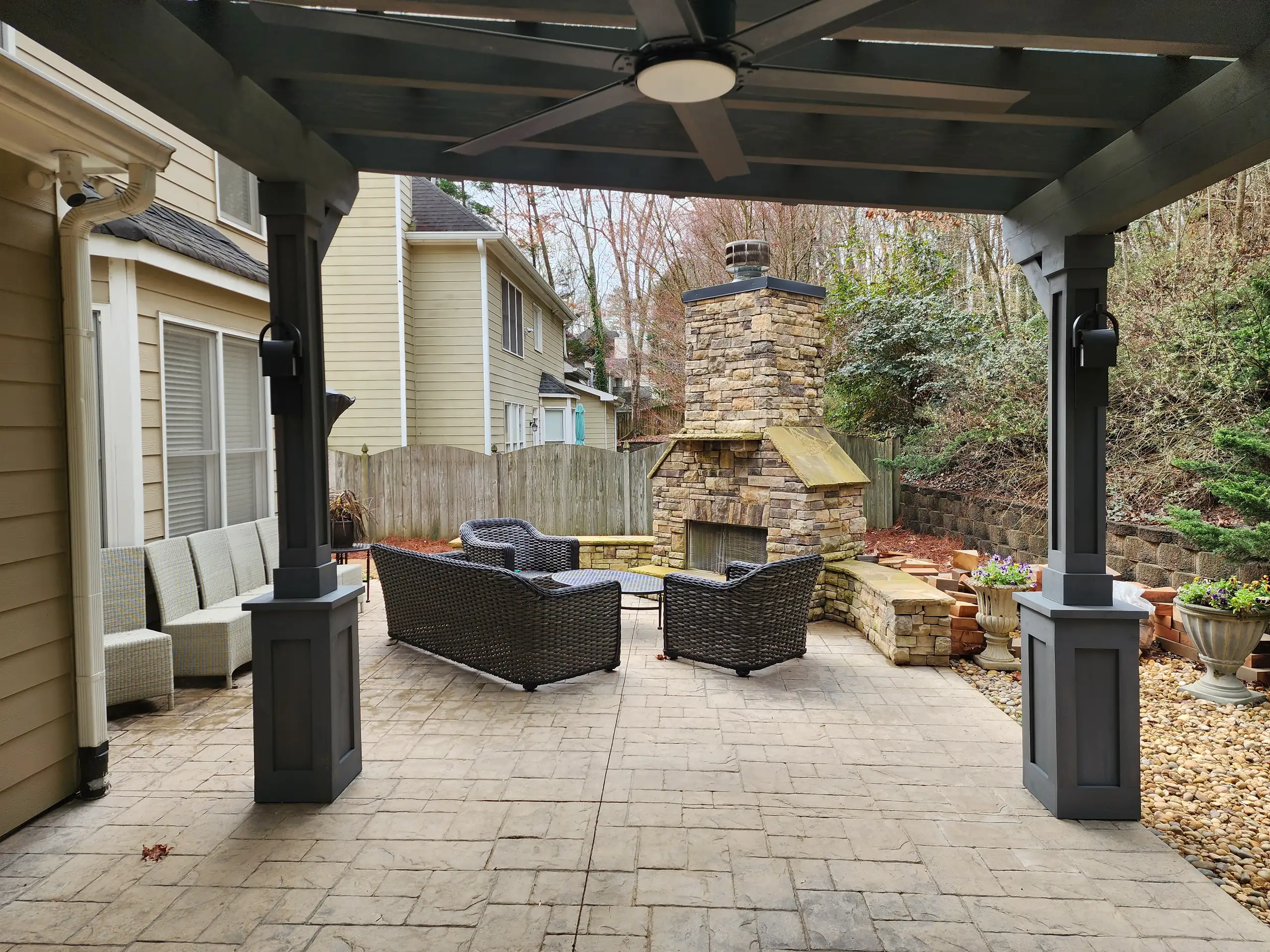outdoor fireplace cost guide Covered patio with dark-painted pergola and ceiling fan, stamped concrete pavers, stone outdoor fireplace and woven wicker seating
